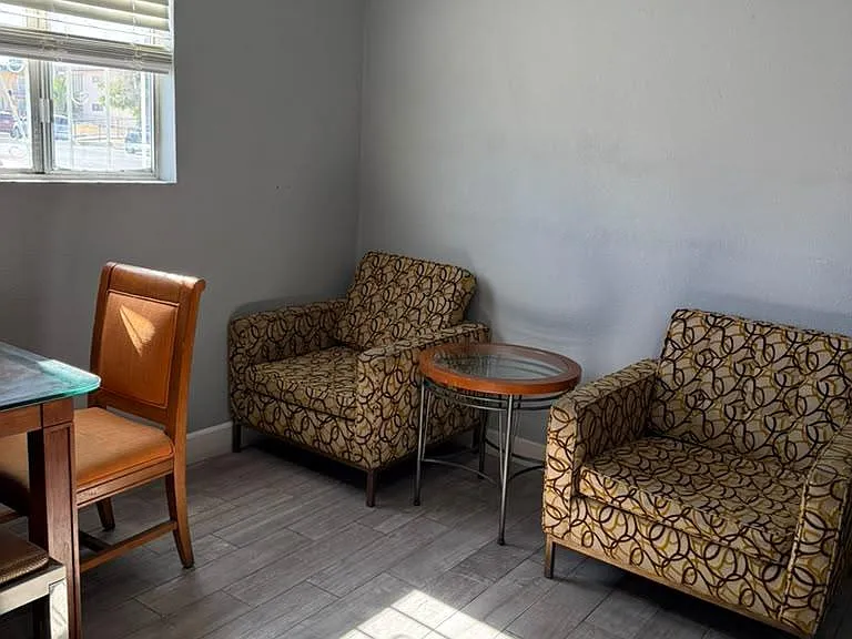 Compact sitting area with two accent chairs, a side table, and a window with blinds over durable tile flooring.