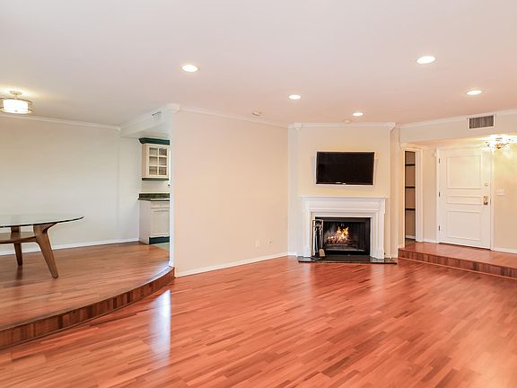 Living Room- Pictured from left to right: Step up dining area, left corner of the Custom Kitchen, fireplace, large wall mounted Flat Screen T.V., doorway leading to Custom Kitchen, front door that leads to the hallway.