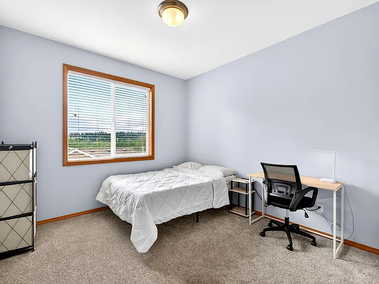 Carpeted bedroom with large window, comfortable bed, and a dedicated desk setup for work or study.