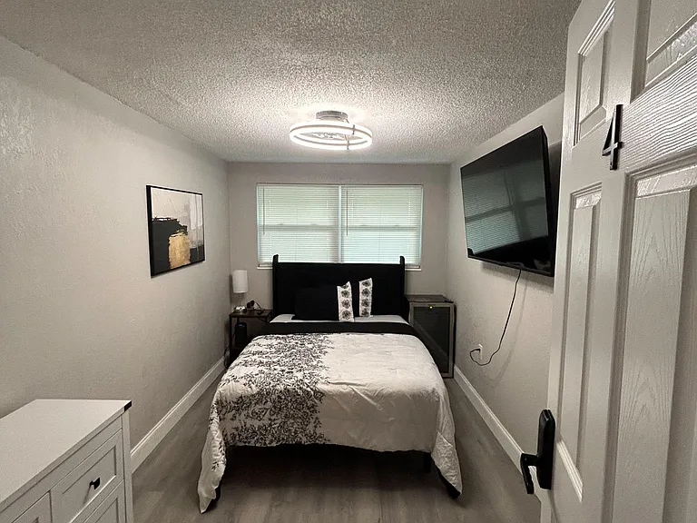 Private bedroom with a full bed, dresser storage, nightstand, and a wall-mounted TV plus a window with blinds.