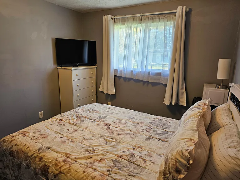 Furnished bedroom with a large window and layered curtains, plus a dresser and TV for convenient everyday living.