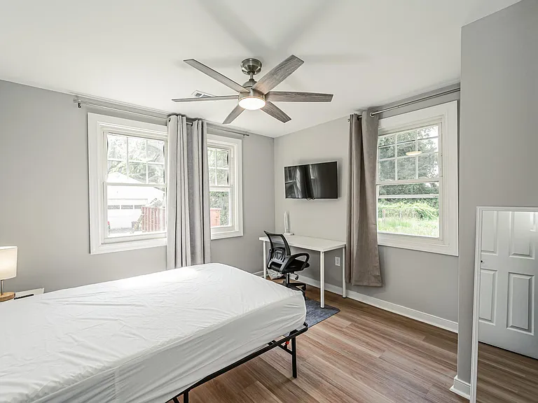Private bedroom with two windows, ceiling fan, wall-mounted TV, and a dedicated desk area for work or study.