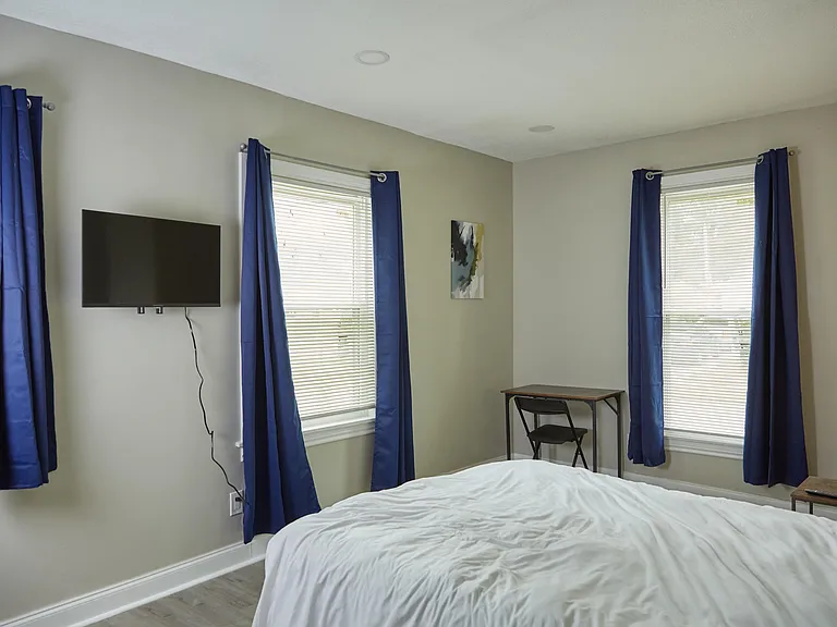 Private bedroom with multiple windows, blackout-style curtains, and a wall-mounted TV plus a compact desk area.