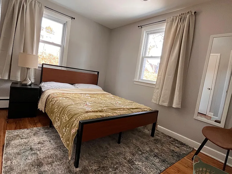 Furnished bedroom with hardwood floors, area rug, two windows with curtains, and a bedside table for essentials.