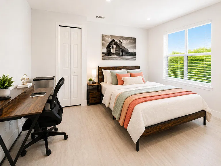 Furnished bedroom with a full bed, nightstand lighting, and a dedicated desk area, plus a wide window and closet storage.