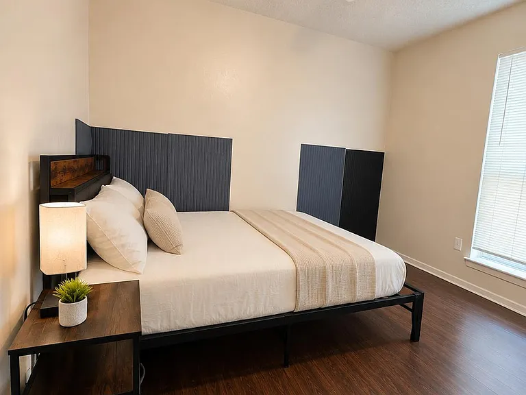 Furnished bedroom with wood-look flooring, ceiling fan with light, and a large window with blinds for privacy.