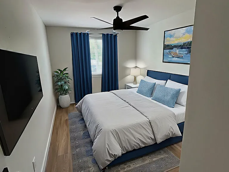 Elegant bedroom with cozy bedding, modern art, and natural light streaming through the window. Features sleek wooden flooring, a large ceiling fan, and stylish blue accents.