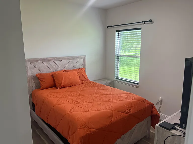 Furnished bedroom featuring a full bed, nightstand, and a large window with blinds for natural light and privacy.