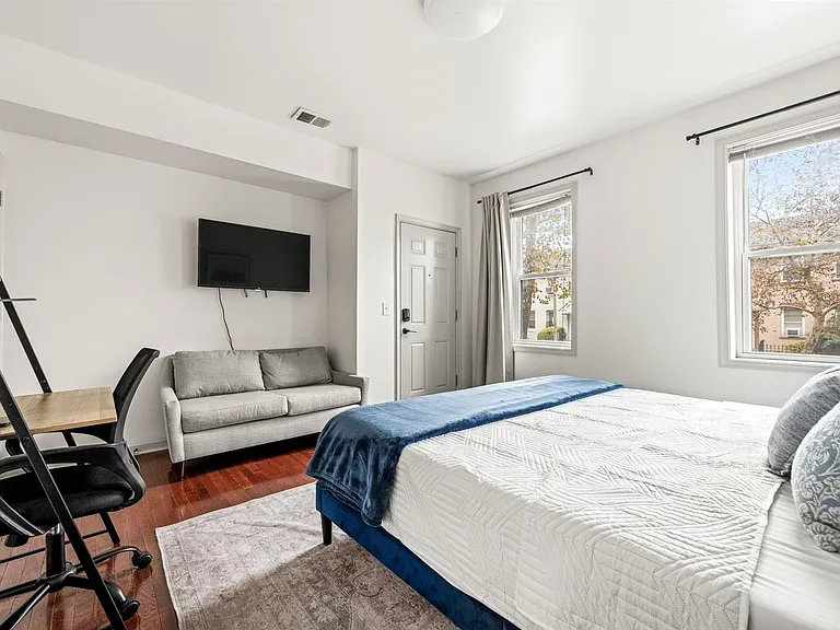 Furnished bedroom featuring a full bed, work desk, loveseat, and wall-mounted TV, plus large windows with curtains.