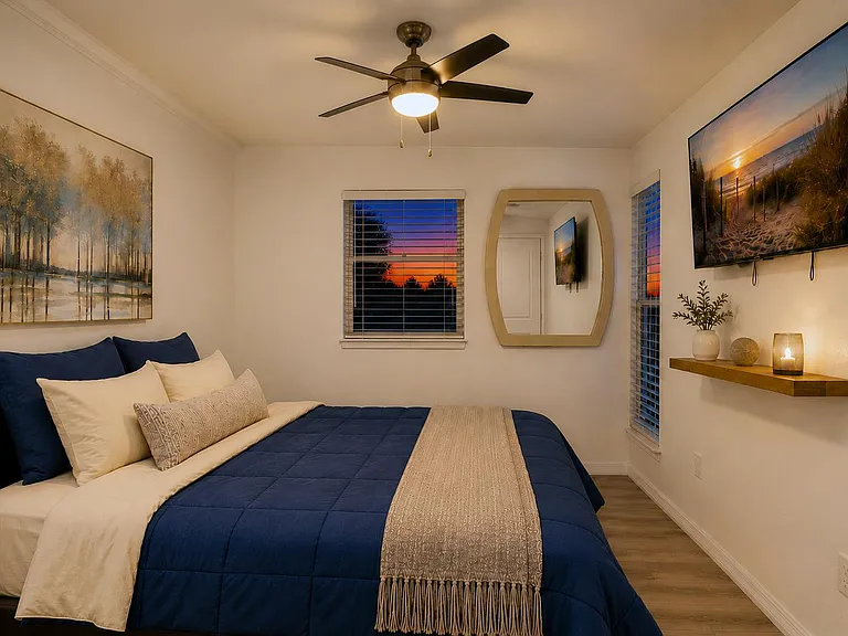 Furnished bedroom with a queen bed, ceiling fan, wall-mounted TV, and windows with blinds for added privacy.