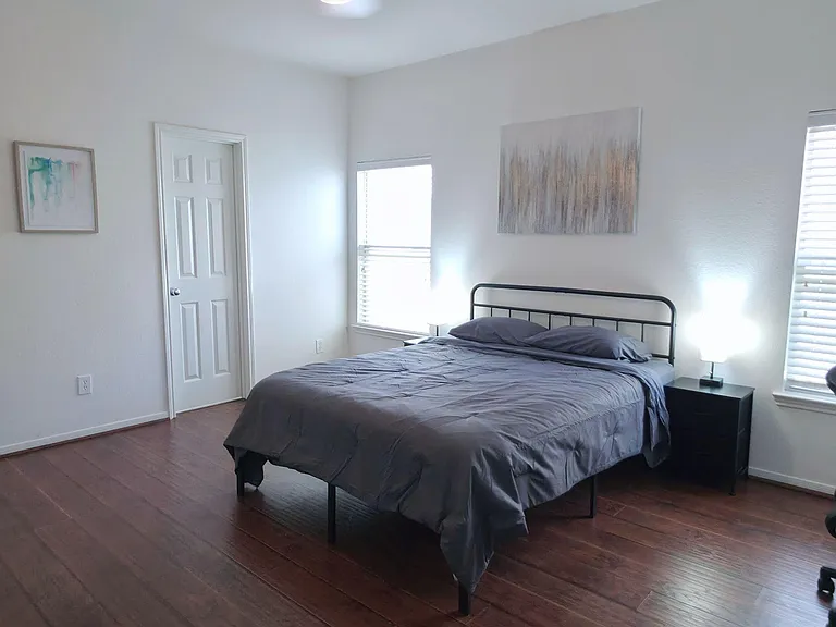 Spacious bedroom with hardwood floors, ceiling fan light, and two windows for natural light plus a bedside table.