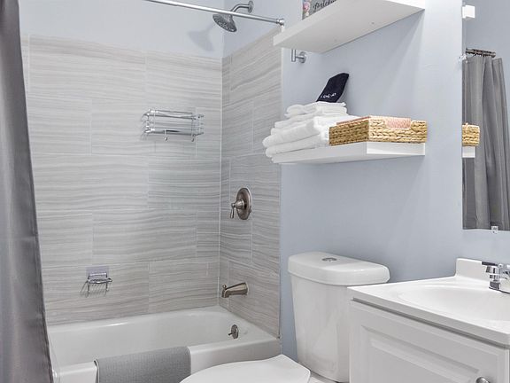 Clean full bath with tiled tub/shower surround, wide vanity with mirror, and open shelving for towels and essentials.