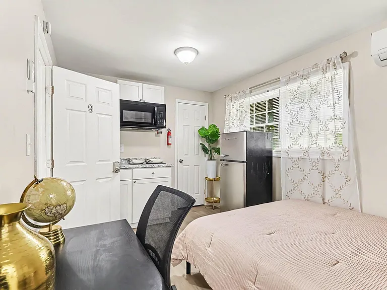 Furnished bedroom with a work desk, mini fridge, and convenient kitchenette storage plus a window and in-room A/C.