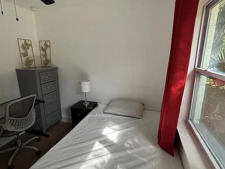 Furnished bedroom with a window, ceiling fan, bed, nightstand with lamp, dresser, and a desk chair for work or study.