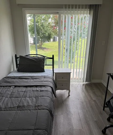 Private bedroom with a full bed, nightstand, and desk, plus sliding glass doors opening to a grassy outdoor view.