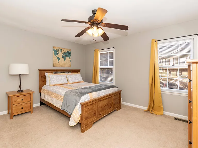 Furnished bedroom with wood bed frame, nightstand and lamp, ceiling fan, and two windows with blinds and curtains.