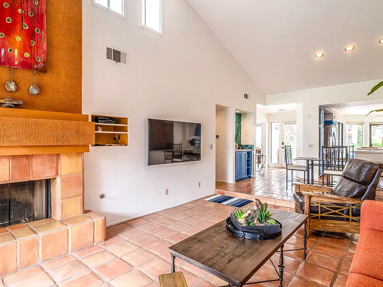 Spacious living room with soaring ceilings, Saltillo tile floors, fireplace, and wall-mounted TV. Sliding doors open to the private greenbelt patio, creating a bright, airy indoor-outdoor flow ideal for relaxed desert living.