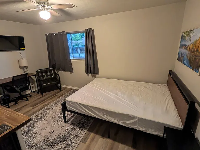 Furnished bedroom with a ceiling fan, window with curtains, and space for a desk setup alongside the bed.