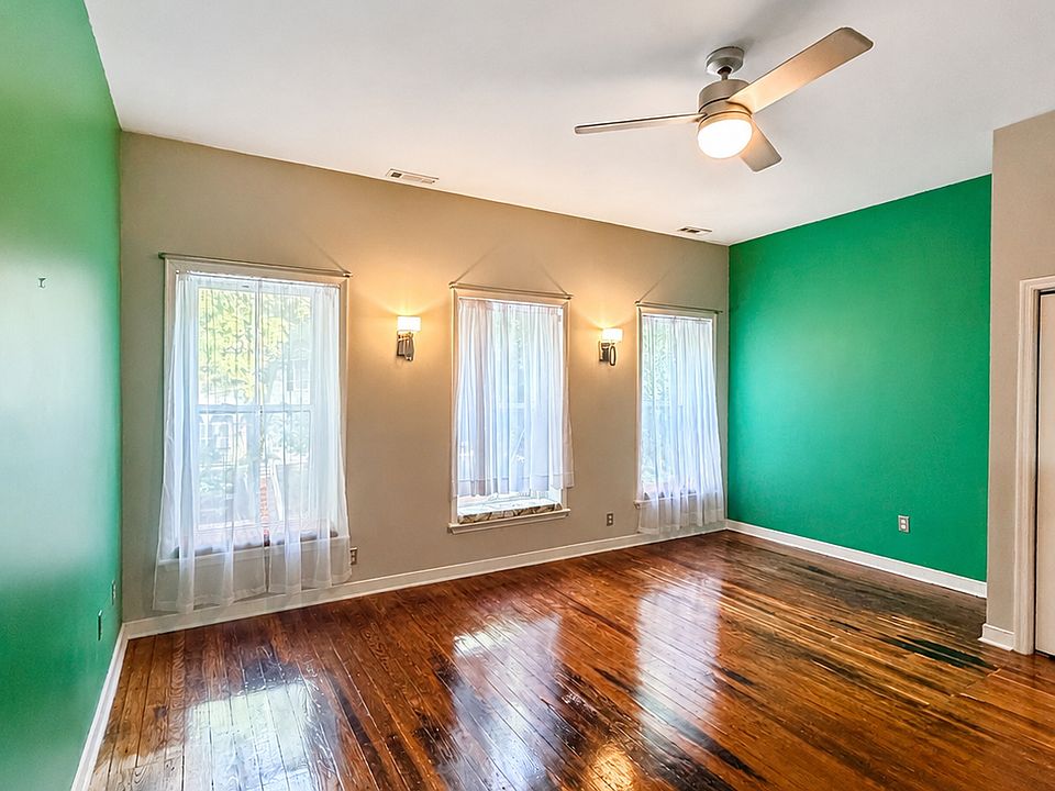 Spacious bedroom with high ceilings, hardwood floors, three large windows, closet, ceiling fan, and warm wall sconces. Bold green accent walls add personality, while the natural light and historic wood floors make the room feel bright and inviting.