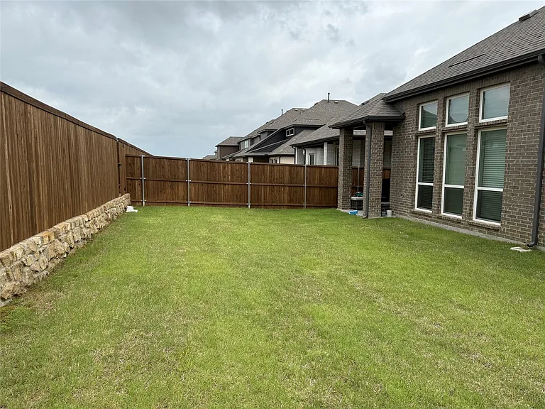 Expansive grassy backyard featuring a full privacy fence, stone retaining wall, and a brick exterior with large windows