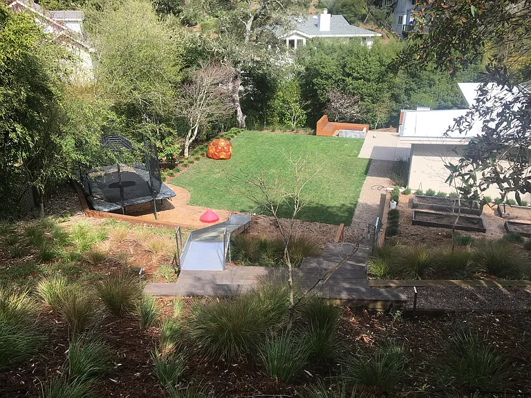 View of private and expansive backyard with slide built into the hillside of native grasses and a trampoline fur jumping.
