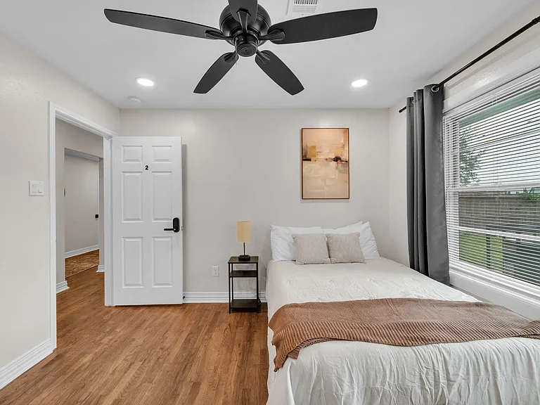 Well-appointed bedroom with wood-look floors, ceiling fan, and a large window with blinds and curtains for natural light.