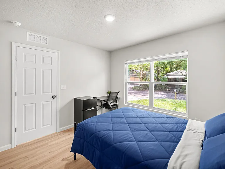 Private bedroom with a large window, wood-look flooring, and space for a desk, plus an in-room mini fridge.
