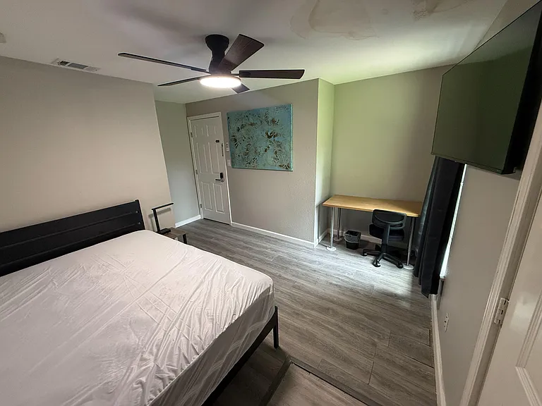 Furnished bedroom with ceiling fan, wall-mounted TV, and a dedicated desk area beside a window with curtains.