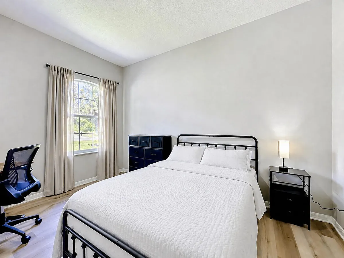 Spacious bedroom featuring a queen bed, nightstand with lamp, dresser storage, and a large window with curtains. - Renshaw Dr
