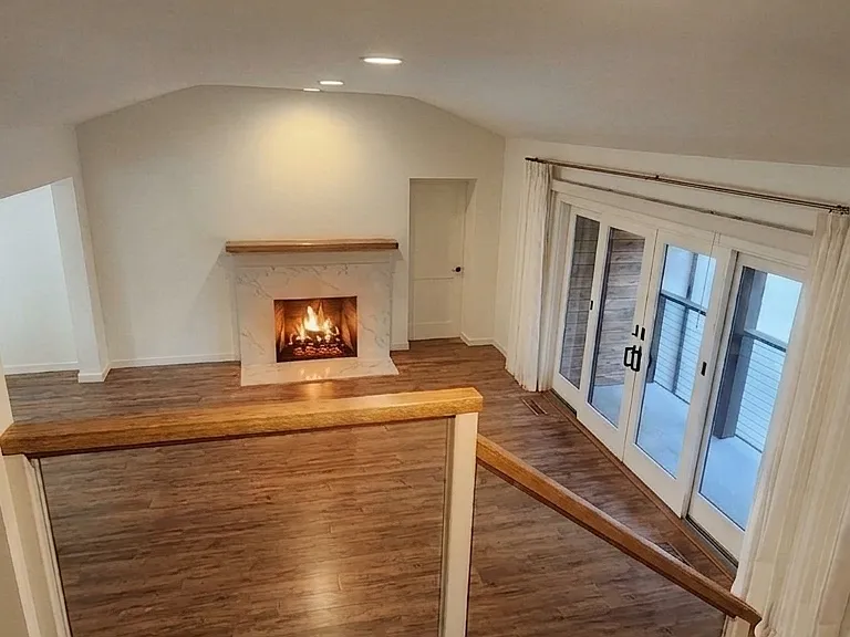 Living room, east-facing view through glass railing from upper level. Gas fireplace, street-side window shades open.
