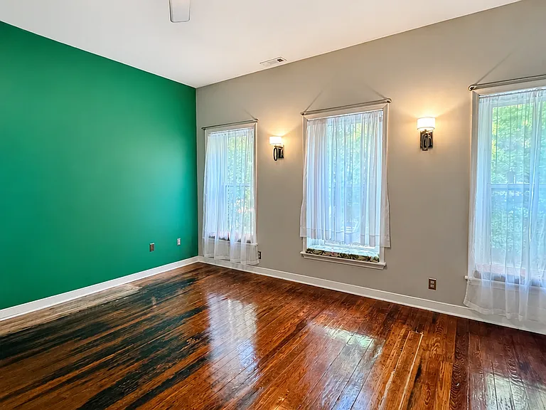 Spacious bedroom with high ceilings, hardwood floors, three large windows, closet, ceiling fan, and warm wall sconces. Bold green accent walls add personality, while the natural light and historic wood floors make the room feel bright and inviting.