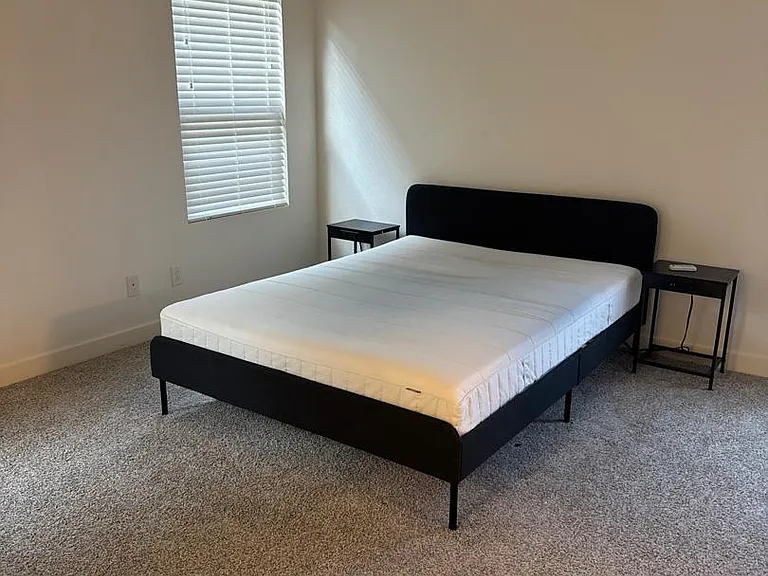 Carpeted bedroom with a queen bed, two nightstands, window blinds, and a ceiling fan for added comfort.