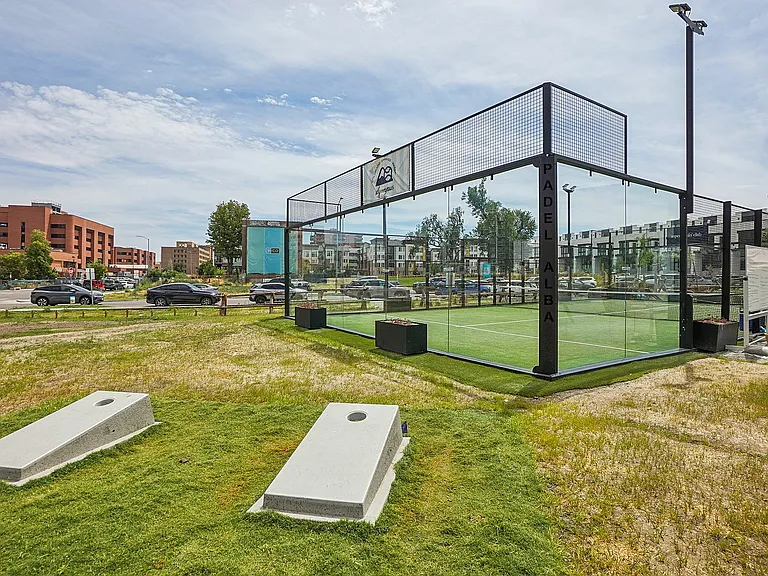 Vibrant outdoor padel court with clear glass walls in a lively urban setting, surrounded by modern buildings and green space.