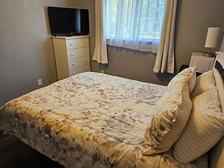 Furnished bedroom with a full bed, dresser with TV, and a window with curtains for natural light and privacy.