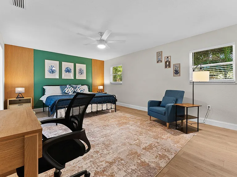 Spacious furnished bedroom with a queen bed, work desk, and seating area, plus ceiling fan and window blinds.