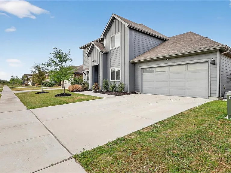 Contemporary gray siding exterior with a contrasting dark front door, attached garage with frosted glass panels, concrete driveway, and a manicured lawn with young trees