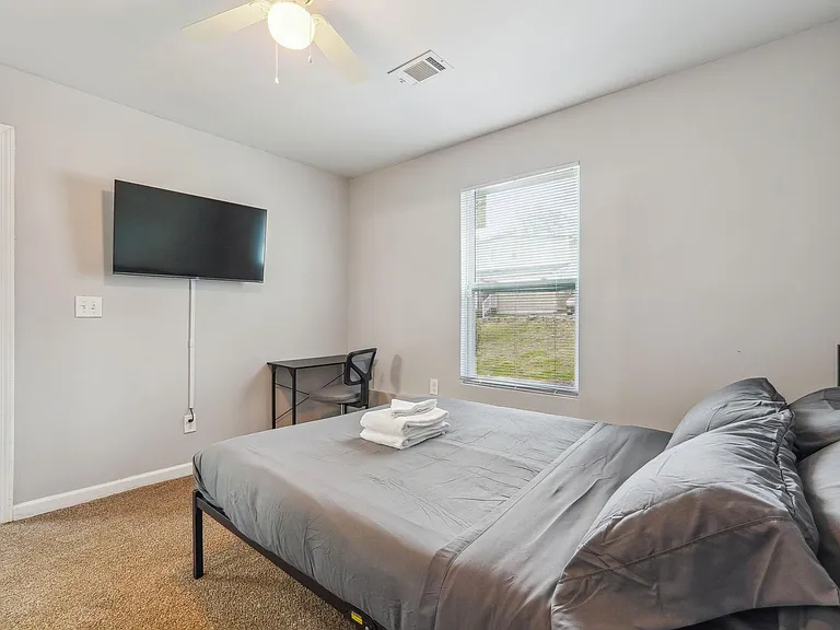 Furnished bedroom with a full bed, wall-mounted TV, and a dedicated desk area, plus a window with blinds for natural light.