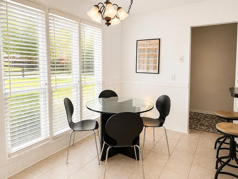 Sunny dining nook with a glass-top table, chandelier lighting, and large windows with shutters overlooking the yard.