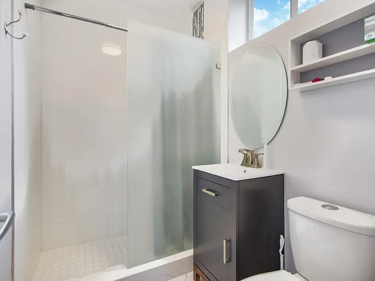Streamlined bathroom with a glass-enclosed shower, single-sink vanity, wall mirror, and open shelving for essentials.