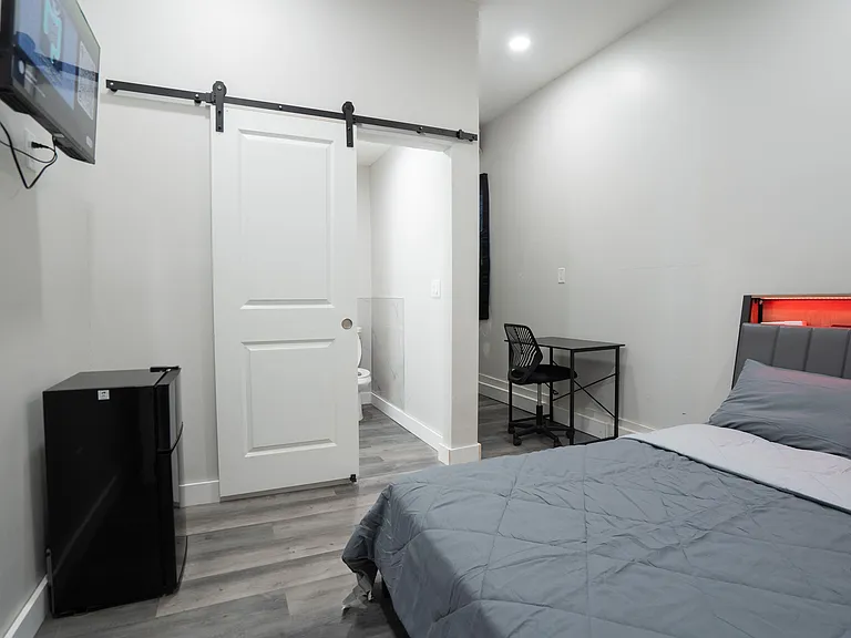 Furnished bedroom with a full bed, wall-mounted TV, mini fridge, and a compact desk workspace plus sliding-door access.