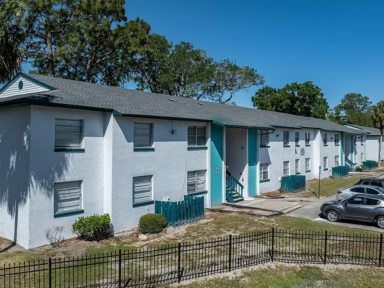 Charming suburban apartment complex with white stucco exterior and teal accents, surrounded by lush trees and ample parking space.