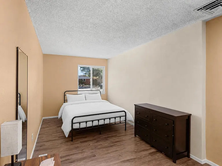 Furnished bedroom with a full bed, dresser, and large mirror, plus a window for natural light and wood-style flooring.
