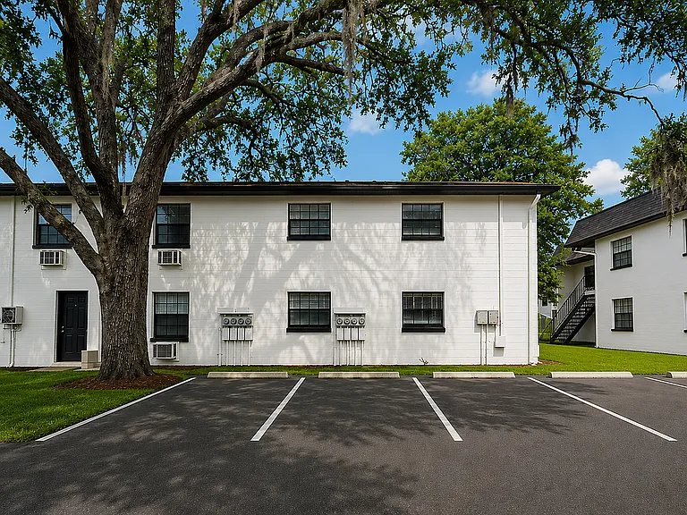 Charming apartment building with a classic white facade, nestled under a beautiful large tree, offering ample parking space in a serene neighborhood.