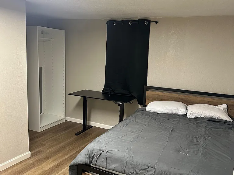 Furnished bedroom with a full bed, wood-style floors, and an open wardrobe plus desk space for work or study.