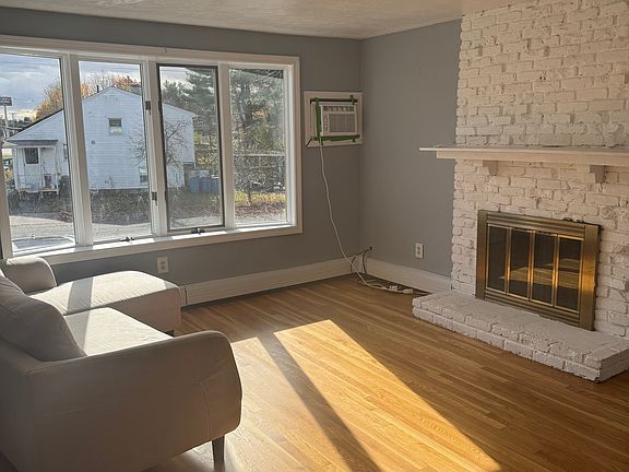 Bright living room featuring refinished hardwood floors, a painted white brick fireplace with mantel, and soft gray walls. Large window brings in excellent natural light.