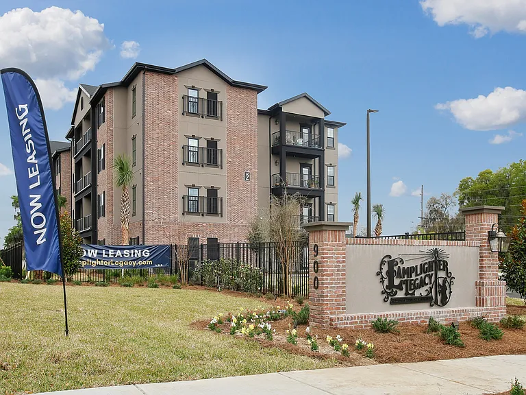 Charming brick apartment complex with modern balconies, welcoming landscaping, and vibrant 'Now Leasing' banners.