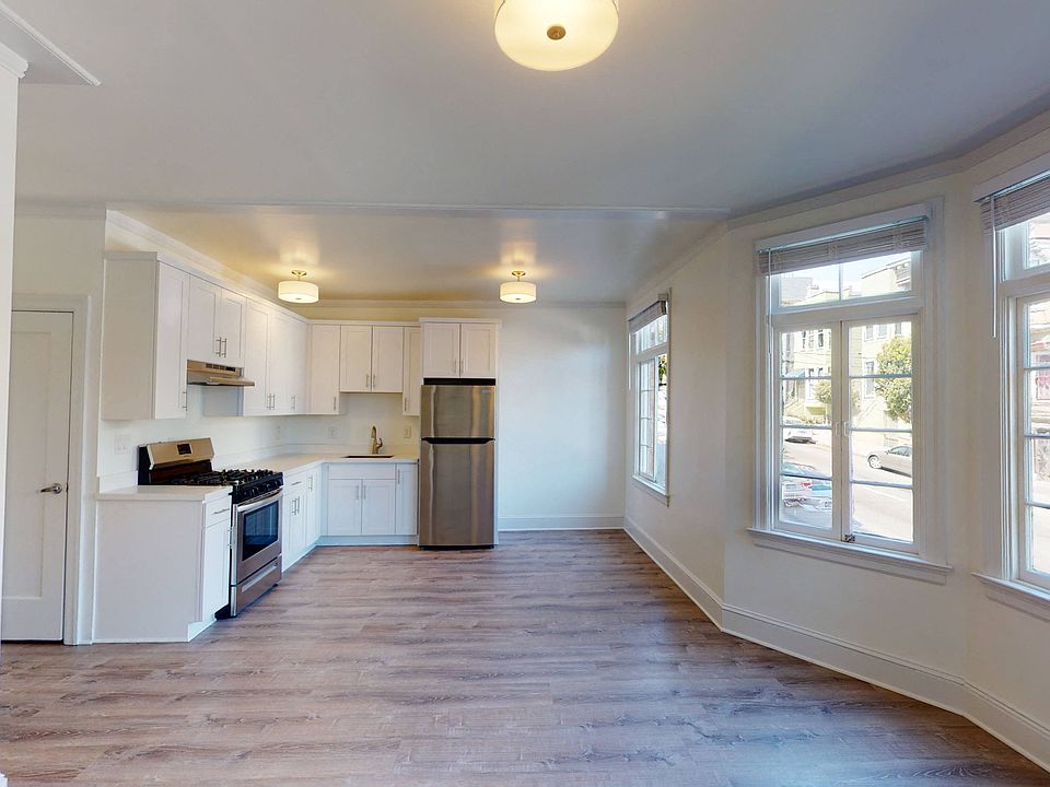 Sunlit living room with wood-like floors, large windows, and a view of the kitchen