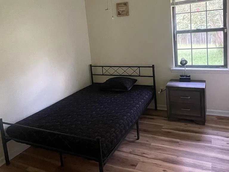Simple furnished bedroom with a twin bed, nightstand, ceiling fan, and window overlooking the yard.
