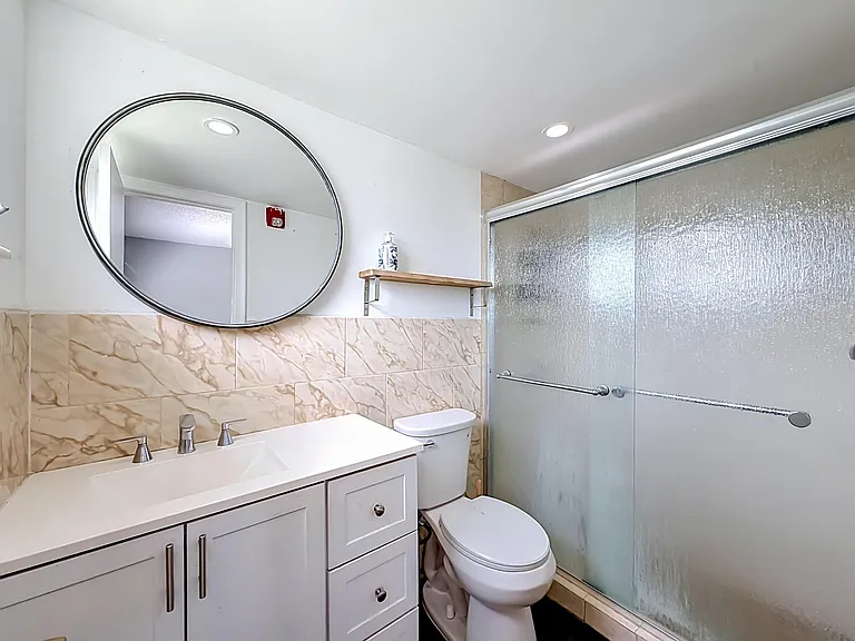 Bathroom with a wide vanity, round mirror, wall shelf, and sliding glass shower doors with tiled surround.
