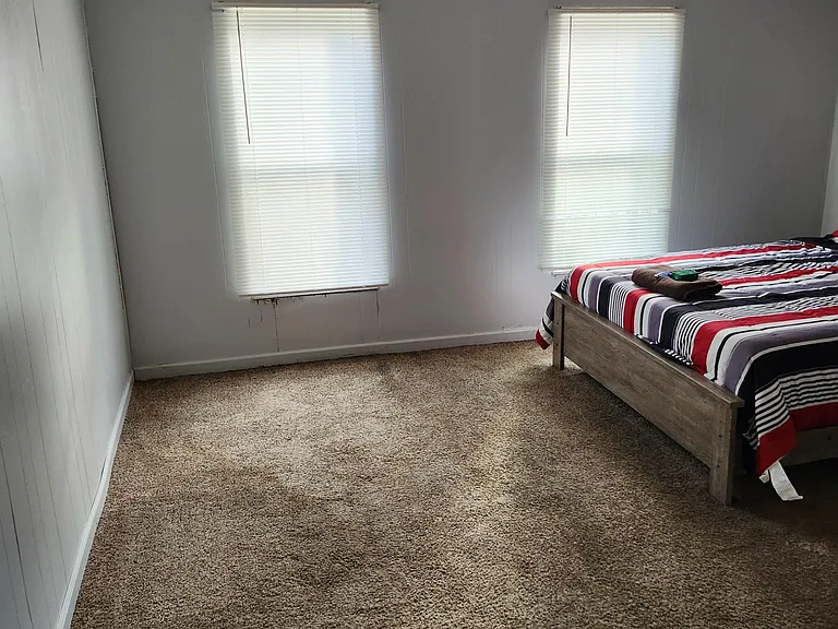 Carpeted bedroom with two windows and blinds, offering flexible space for a full/queen bed and a small dresser.